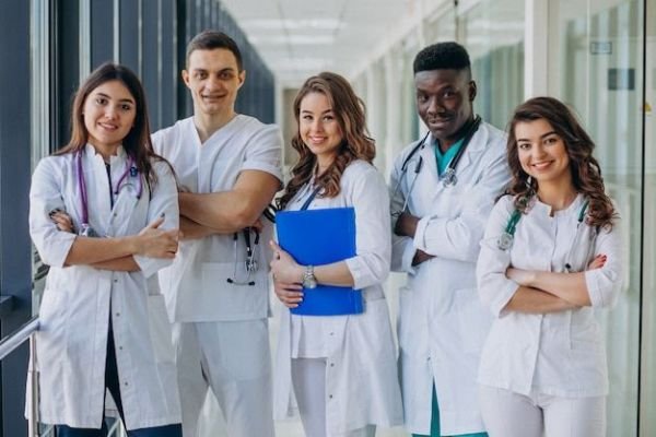 Group of five healthcare professionals in white coats standing in a bright hospital corridor, smiling at the camera; one holds a blue clipboard.